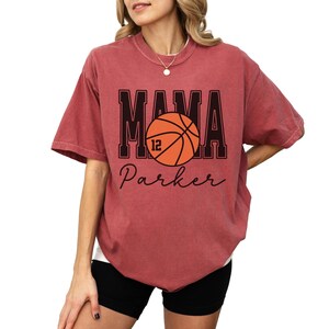 custom basketball mom shirt. the basketball shirt says mama in large varsity letters. there is a large basketball in the middle that is customized with the players number. the basketball tshirt is personalized with the players name below the basketball in a script font. comfort colors basketball mama shirt, short sleeves, crewneck tee