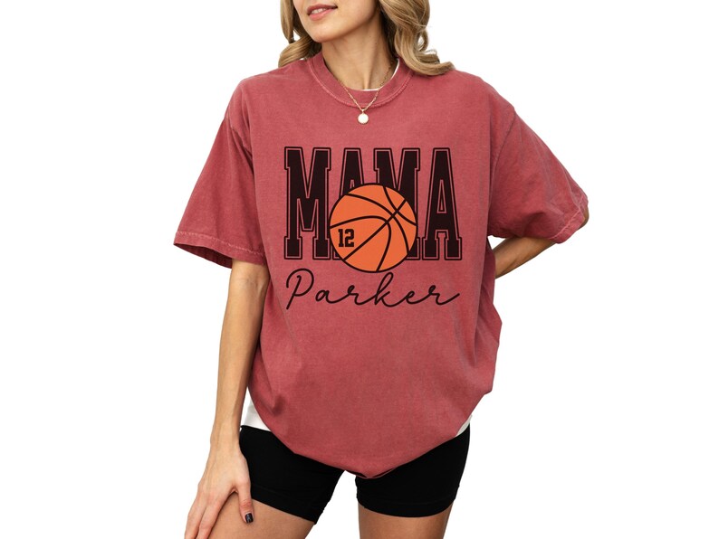 custom basketball mom shirt. the basketball shirt says mama in large varsity letters. there is a large basketball in the middle that is customized with the players number. the basketball tshirt is personalized with the players name below the basketball in a script font. comfort colors basketball mama shirt, short sleeves, crewneck tee
