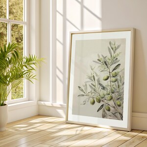 May include: A framed print depicting an olive branch with green olives and foliage. The artwork is in a light wood frame, leaning against a white wall. The room features a wooden floor and a potted plant in the corner.