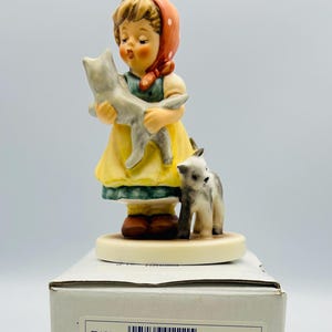 May include: A porcelain figurine of a girl in a yellow dress and red headscarf holding a gray cat, with another cat at her feet. The figurine is on a round base, with a box and certificate in the background. The box has the text "Tender Love #1111".