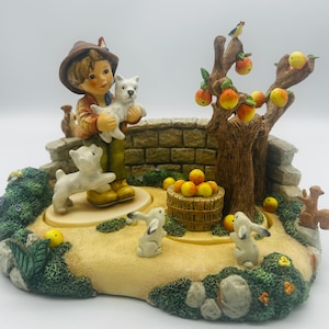 May include: A detailed figurine depicting a young boy holding a white puppy, surrounded by a miniature garden scene. The scene includes a tree with apples, a stone bridge, rabbits, and a squirrel. The color palette is primarily earth tones.