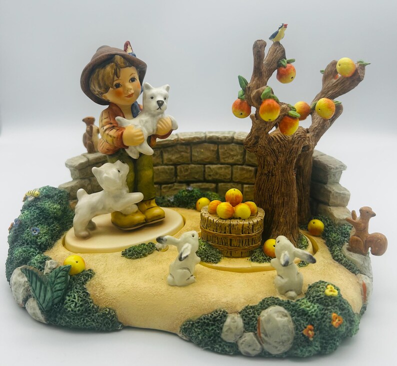 May include: A detailed figurine depicting a young boy holding a white puppy, surrounded by a miniature garden scene. The scene includes a tree with apples, a stone bridge, rabbits, and a squirrel. The color palette is primarily earth tones.