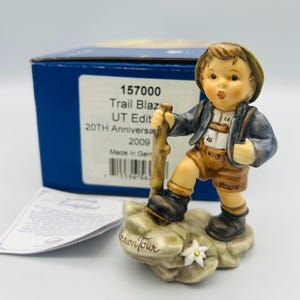 Może przedstawiać: Porcelanowa figurka młodego turysty z laską, ubranego w niebieską kurtkę, brązowe szorty i zielony kapelusz. Figurka stoi na skale z kwiatem. Na pudełku napis "Trail Blazer" i "20th Anniversary 2009".