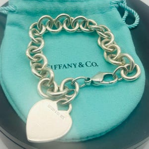 May include: A sterling silver chain link bracelet featuring a heart-shaped charm. The charm is engraved with the text Tiffany & Co. 925. The bracelet includes a lobster claw clasp and is presented on a light blue fabric drawstring pouch.