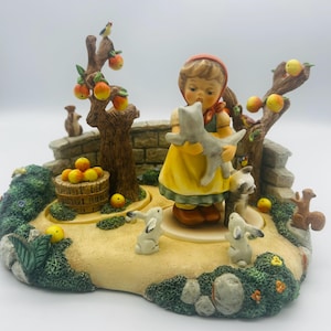 May include: A detailed figurine depicting a young girl in a yellow dress holding a gray cat, surrounded by trees with apples, rabbits, squirrels, and a stone wall. The base is a garden scene with a basket of apples.