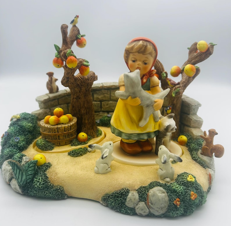 May include: A detailed figurine depicting a young girl in a yellow dress holding a gray cat, surrounded by trees with apples, rabbits, squirrels, and a stone wall. The base is a garden scene with a basket of apples.