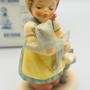 May include: A ceramic figurine of a young girl in a yellow dress and red headscarf, holding a gray cat. Another gray cat stands at her feet. The figurine has a cream-colored base and is painted with soft colors.