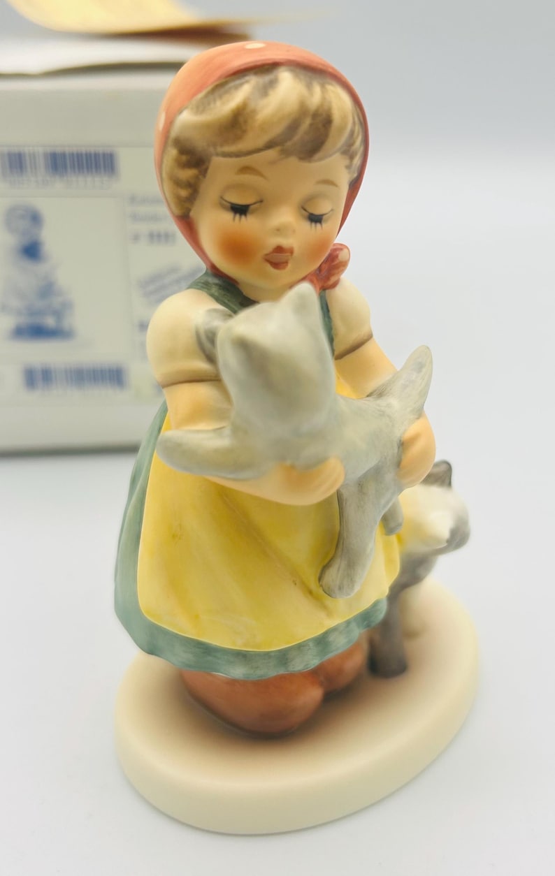 May include: A ceramic figurine of a young girl in a yellow dress and red headscarf, holding a gray cat. Another gray cat stands at her feet. The figurine has a cream-colored base and is painted with soft colors.