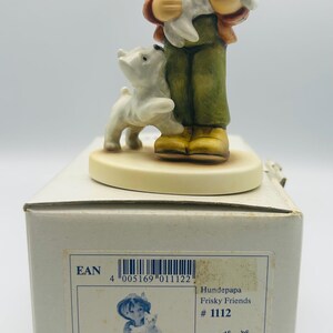 May include: A Hummel figurine of a young person holding a white dog, with another white dog at their feet. The figurine is on a white base, and the box reads "Frisky Friends #1112" and "Hum 2008", with a height of 4 3/8 inches.