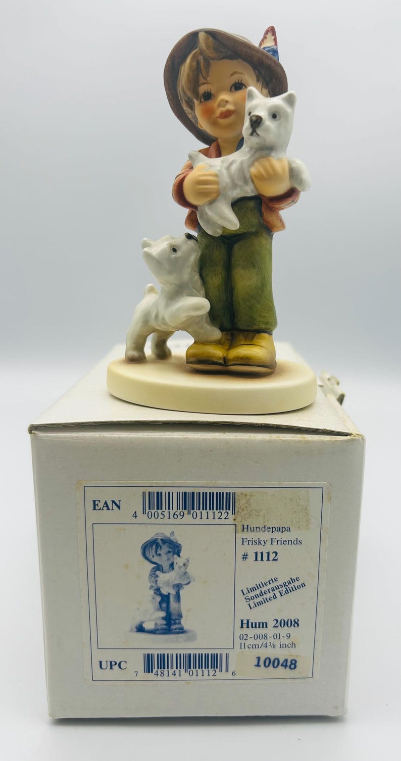 May include: A Hummel figurine of a young person holding a white dog, with another white dog at their feet. The figurine is on a white base, and the box reads "Frisky Friends #1112" and "Hum 2008", with a height of 4 3/8 inches.