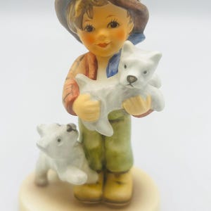 May include: A ceramic figurine of a young person holding two white puppies. The figure wears a brown hat, a red and orange jacket, green pants, and yellow boots. The figurine is on a cream-colored base.