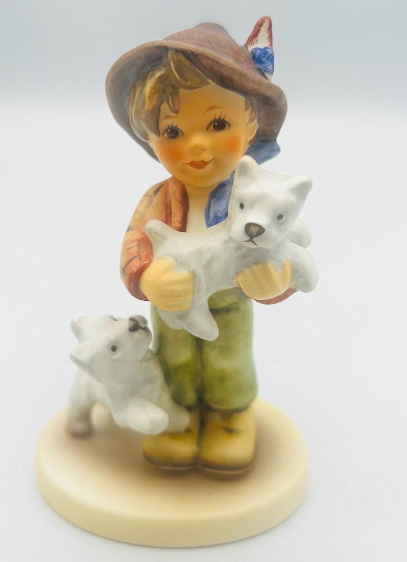 May include: A ceramic figurine of a young person holding two white puppies. The figure wears a brown hat, a red and orange jacket, green pants, and yellow boots. The figurine is on a cream-colored base.