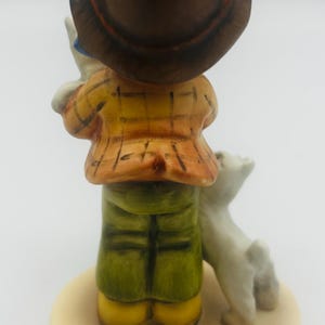 May include: A ceramic figurine of a person in a yellow plaid jacket, green pants, and a brown hat, holding a small white dog. The figurine is on a beige base with the text "Hundepapa Frisky Friends".