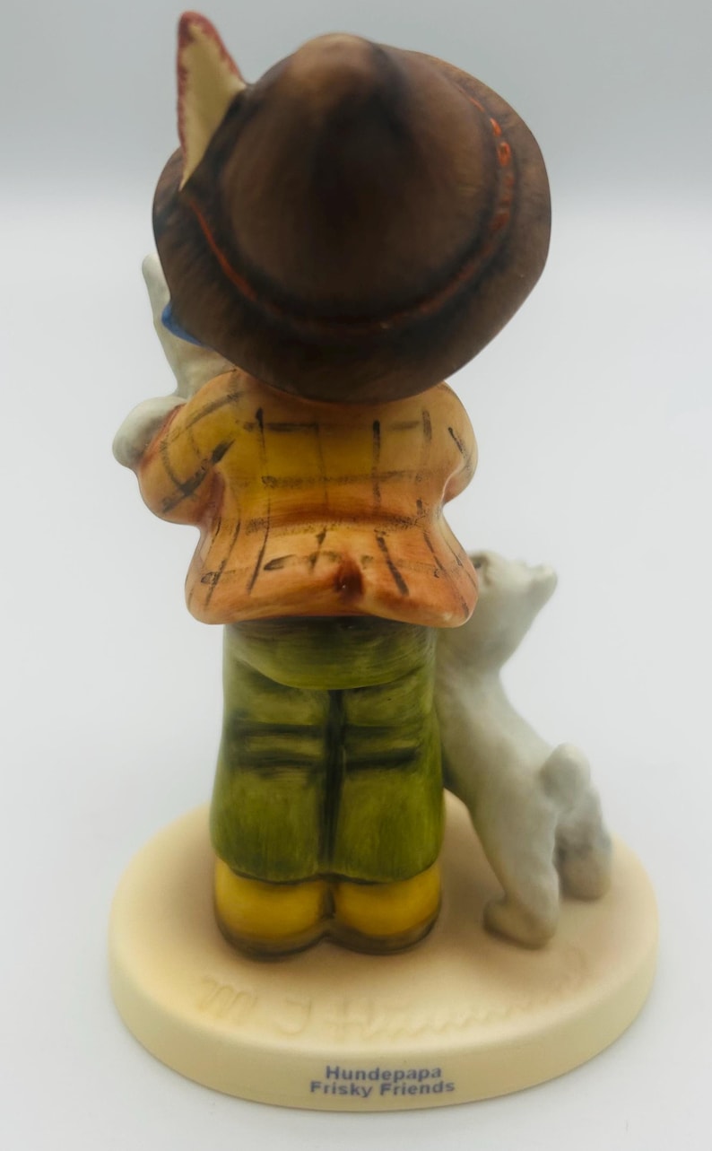May include: A ceramic figurine of a person in a yellow plaid jacket, green pants, and a brown hat, holding a small white dog. The figurine is on a beige base with the text "Hundepapa Frisky Friends".