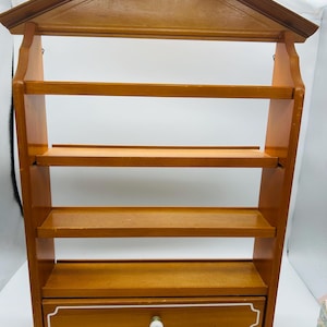 Pu&ograve; includere: Una mensola da parete in legno con una parte superiore a forma di casa e quattro ripiani. Il ripiano inferiore ha un piccolo cassetto con finiture bianche e un pomello rotondo bianco. Il legno ha un caldo tono marrone medio.