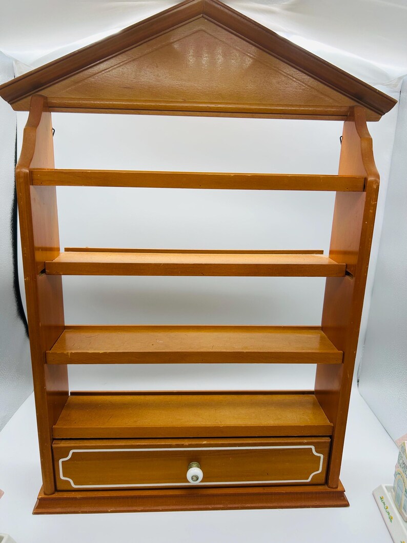 Pu&ograve; includere: Una mensola da parete in legno con una parte superiore a forma di casa e quattro ripiani. Il ripiano inferiore ha un piccolo cassetto con finiture bianche e un pomello rotondo bianco. Il legno ha un caldo tono marrone medio.