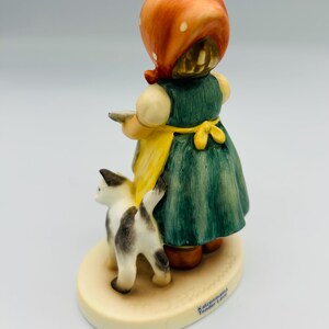 May include: A ceramic figurine of a girl in a green dress and yellow apron, wearing an orange headscarf. A black and white cat stands beside her. The figurine is on a cream-colored base with the words "Katzenmama Tender Love".
