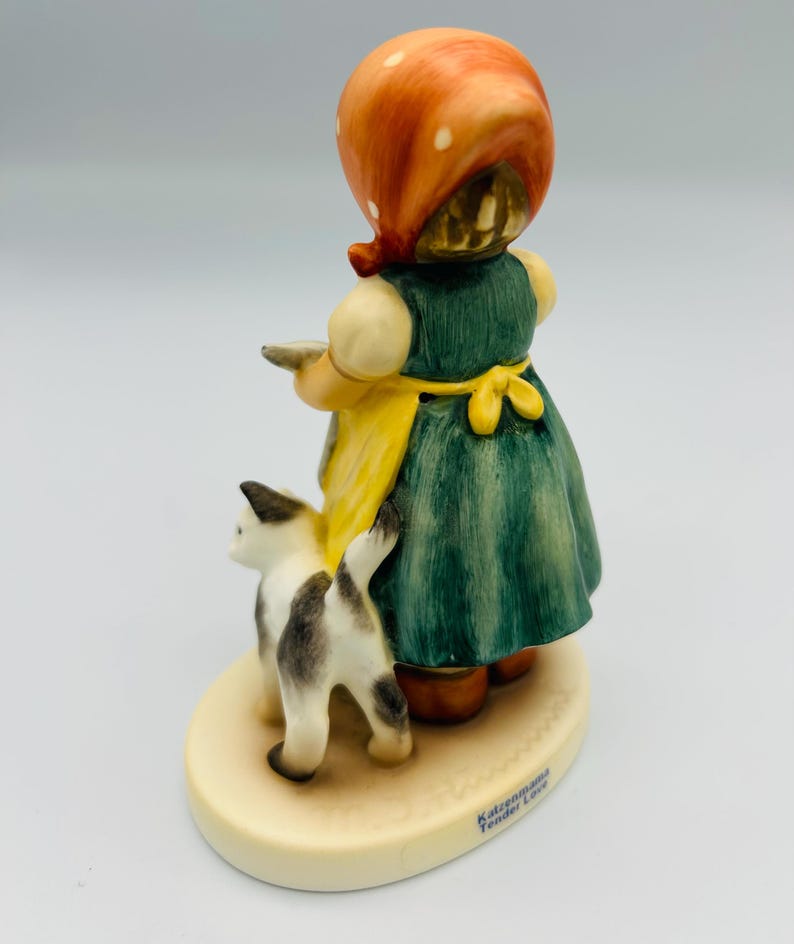 May include: A ceramic figurine of a girl in a green dress and yellow apron, wearing an orange headscarf. A black and white cat stands beside her. The figurine is on a cream-colored base with the words "Katzenmama Tender Love".