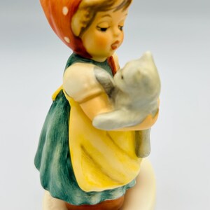 May include: A ceramic figurine of a young girl holding a gray cat. The girl wears a red headscarf with white polka dots, a green dress, and a yellow apron. The figurine is on a cream-colored base.