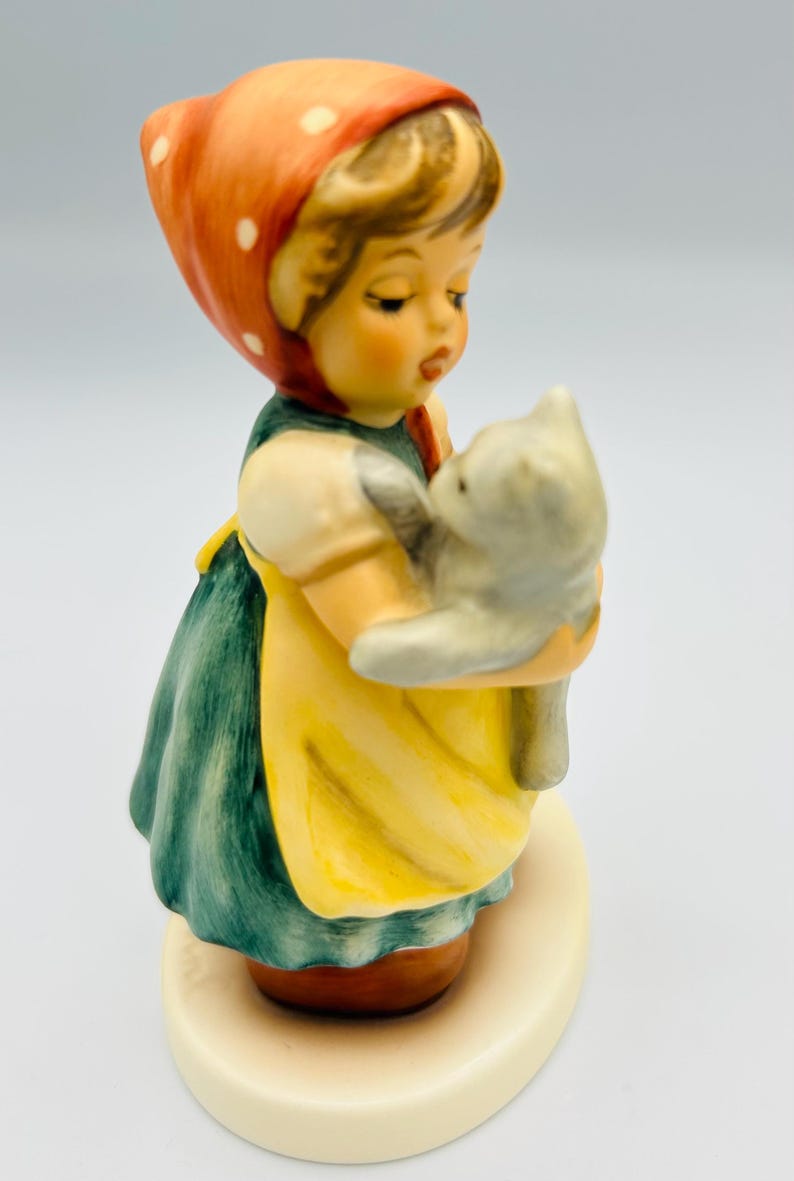 May include: A ceramic figurine of a young girl holding a gray cat. The girl wears a red headscarf with white polka dots, a green dress, and a yellow apron. The figurine is on a cream-colored base.