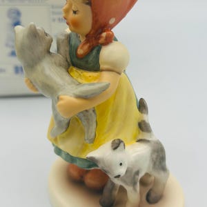 May include: A ceramic figurine of a young girl in a yellow dress and red headscarf, holding a gray cat. Another gray and white cat stands at her feet. The figurine is on a cream-colored base.