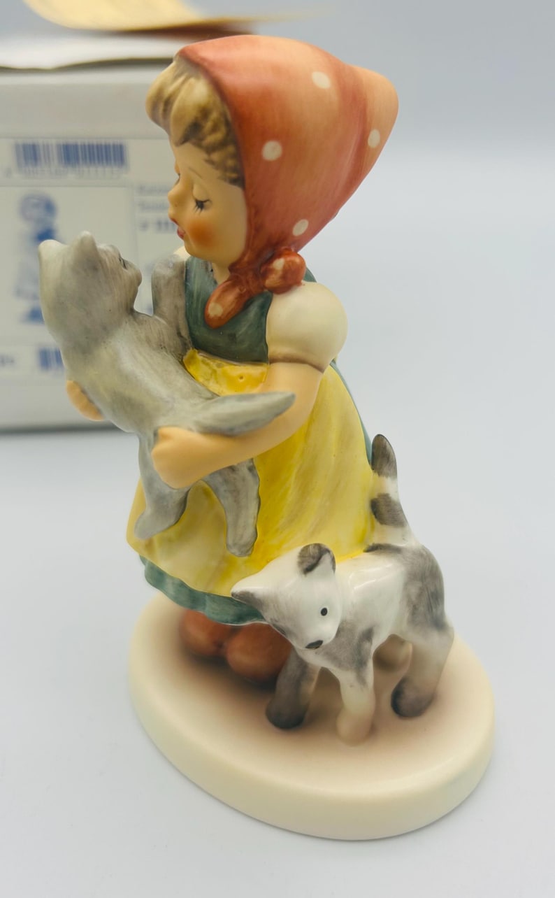 May include: A ceramic figurine of a young girl in a yellow dress and red headscarf, holding a gray cat. Another gray and white cat stands at her feet. The figurine is on a cream-colored base.