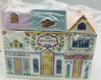 May include: A ceramic recipe box in the shape of a house, featuring pastel colors and floral accents. The box has multiple sections, with the word "RECIPES" displayed prominently. The design includes windows, doors, and a roof, all in a charming, decorative style.