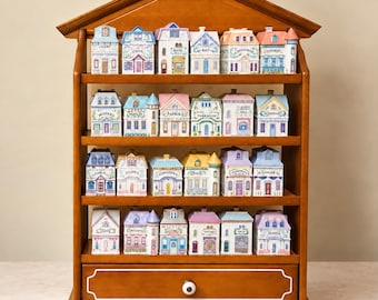 Op de afbeelding: Een houten displayplank in de vorm van een huis met vier niveaus, gevuld met vierentwintig keramische kruidenpotjes ontworpen als miniatuurhuisjes. Elk potje heeft een unieke bouwstijl, pastelkleuren en een specifieke kruidnaam op de voorzijde.