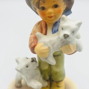 May include: A ceramic figurine of a young person holding a white puppy, with another puppy at their feet. The figurine has a brown hat, a red jacket, green pants, and yellow boots. The base is a light beige color.