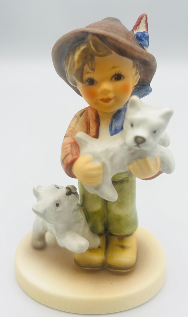 May include: A ceramic figurine of a young person holding a white puppy, with another puppy at their feet. The figurine has a brown hat, a red jacket, green pants, and yellow boots. The base is a light beige color.