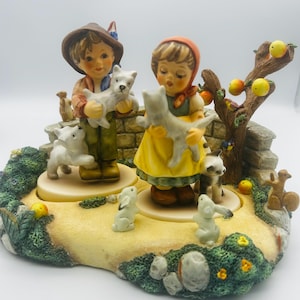 May include: A detailed ceramic figurine depicting two children holding puppies, surrounded by other animals and a tree with apples. The children are dressed in traditional clothing, and the base is decorated with greenery and a stone wall.
