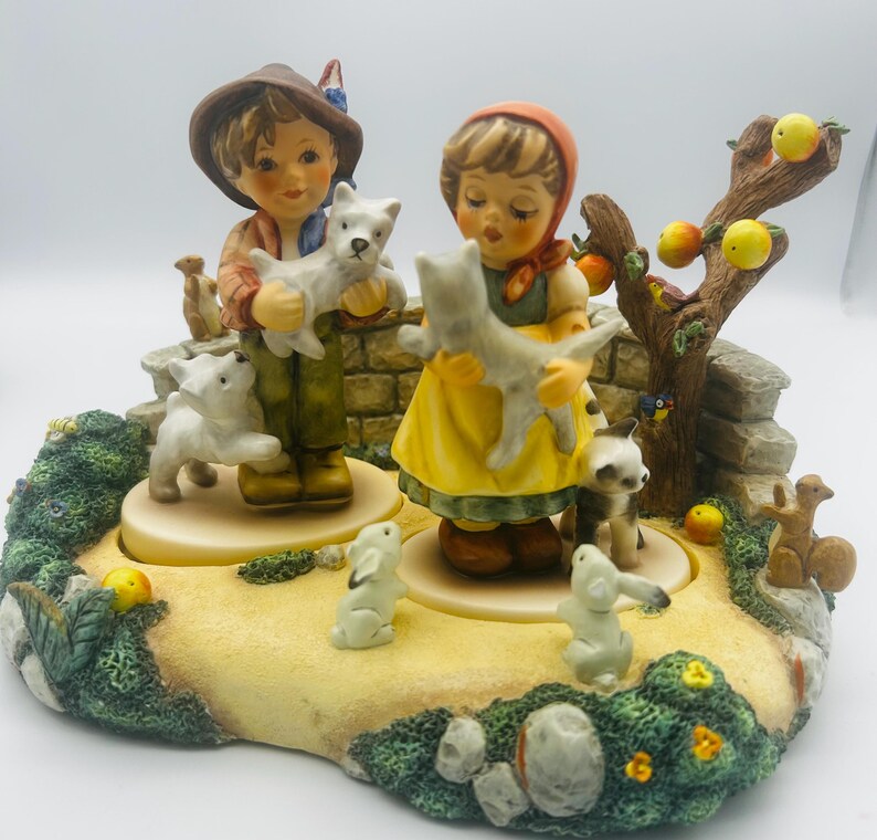 May include: A detailed ceramic figurine depicting two children holding puppies, surrounded by other animals and a tree with apples. The children are dressed in traditional clothing, and the base is decorated with greenery and a stone wall.