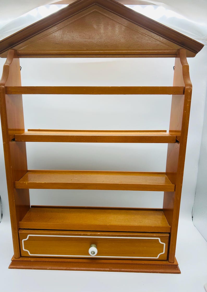 Pu&ograve; includere: Mensola da parete in legno con parte superiore a forma di casa, quattro ripiani e un cassetto con pomello bianco. La mensola &egrave; in legno marrone chiaro.
