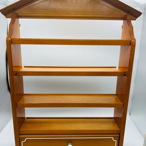 Pu&ograve; includere: Un portapezie in legno da parete con una parte superiore a forma di casa. Presenta quattro ripiani e un cassetto con pomello e finiture bianche. Il portapezie &egrave; realizzato in legno marrone chiaro.