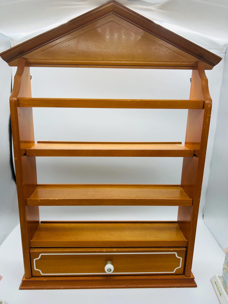 Pu&ograve; includere: Un portapezie in legno da parete con una parte superiore a forma di casa. Presenta quattro ripiani e un cassetto con pomello e finiture bianche. Il portapezie &egrave; realizzato in legno marrone chiaro.
