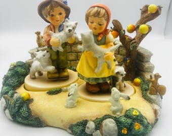 Puede incluir: Figura de cerámica que representa a un niño y una niña sosteniendo cachorros blancos, rodeados de otras figuras de animales. El niño lleva un sombrero marrón y pantalones verdes, mientras que la niña lleva un vestido amarillo. También hay un árbol frutal.