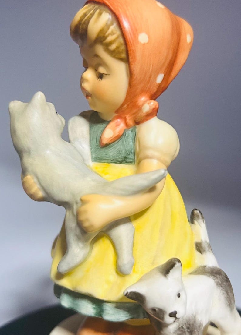 May include: A porcelain figurine of a young girl holding a gray cat, with two additional cats at her feet. The girl wears a yellow dress, green apron, and a red headscarf with white polka dots. The figurine has a vintage aesthetic.