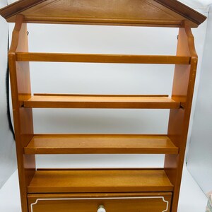 Pu&ograve; includere: Una mensola da parete in legno con una parte superiore a forma di casa e quattro ripiani. Il ripiano inferiore ha un piccolo cassetto con finiture bianche e un pomello rotondo bianco. Il legno ha un caldo tono marrone medio.