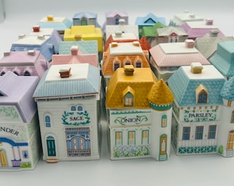 May include: A collection of ceramic spice jars shaped like colorful houses. Each house has a different color roof and trim, with labels like "Coriander," "Sage," and "Onion." The jars are arranged on a white surface.