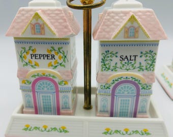 May include: A ceramic salt and pepper set featuring house-shaped shakers with pink roofs and floral accents. The shakers are labeled "PEPPER" and "SALT" and sit on a matching tray with a gold-toned handle. The set is decorated with pastel colors.