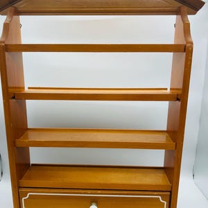 Pu&ograve; includere: Mensola da parete in legno con parte superiore a forma di casa, quattro ripiani e un cassetto. La mensola &egrave; realizzata in legno marrone chiaro con un cassetto bordato di bianco e un pomello rotondo bianco. I ripiani sono larghi circa 30 cm.