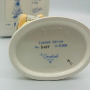 May include: A white ceramic figurine base with the text "2007", "Limited Edition No. 1465 of 25.000", and "Goebel Germany". The figurine is partially visible, with a yellow and gray color scheme.