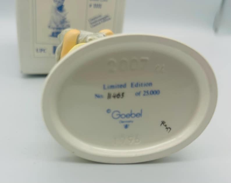 May include: A white ceramic figurine base with the text "2007", "Limited Edition No. 1465 of 25.000", and "Goebel Germany". The figurine is partially visible, with a yellow and gray color scheme.