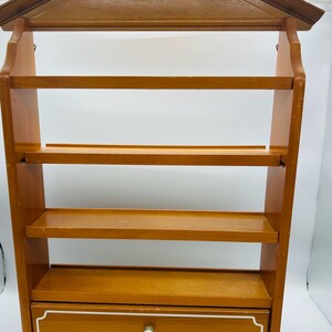 Pu&ograve; includere: Una mensola da parete in legno con una parte superiore a forma di casa e quattro ripiani. Il ripiano inferiore ha un cassetto con un pomello bianco e un bordo bianco decorativo. La mensola &egrave; in legno marrone chiaro.