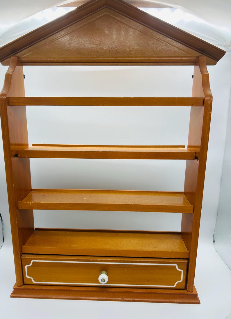 Pu&ograve; includere: Una mensola da parete in legno con una parte superiore a forma di casa e quattro ripiani. Il ripiano inferiore ha un cassetto con un pomello bianco e un bordo bianco decorativo. La mensola &egrave; in legno marrone chiaro.