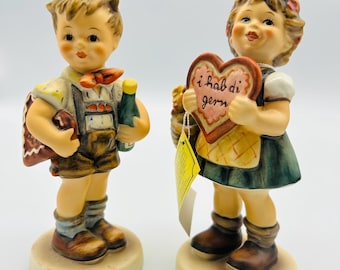 May include: A pair of vintage porcelain figurines of a boy and girl. The boy is holding a bottle and a gingerbread cookie. The girl is holding a heart-shaped cookie with the text "ich hab di gern" on it.