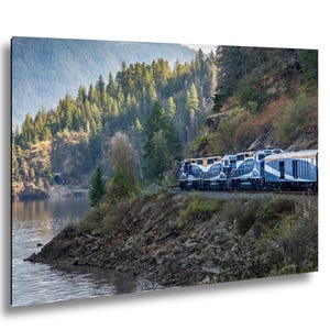 May include: A scenic photograph of a train traveling along a track next to a lake. The train is blue and white, with a mountain range and forest in the background. The water is calm and reflects the surrounding landscape.