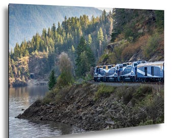May include: A scenic photograph of a train traveling along a track next to a lake. The train is blue and white, with a mountain range and forest in the background. The water is calm and reflects the surrounding landscape.
