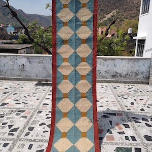 May include: A long rectangular runner rug featuring a repeating geometric pattern of beige diamonds bordered by blue shapes and small mustard yellow squares. The rug is framed by a solid dark red border along both long edges and the top edge.