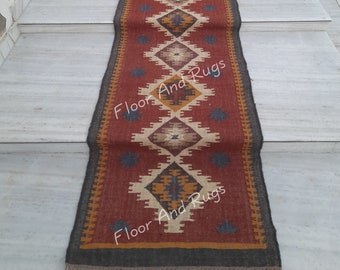 May include: A long, narrow runner rug featuring a deep red base color with a repeating geometric diamond pattern in shades of cream, mustard yellow, and dark blue. The rug has a dark gray border and short, light-colored fringe at both ends.